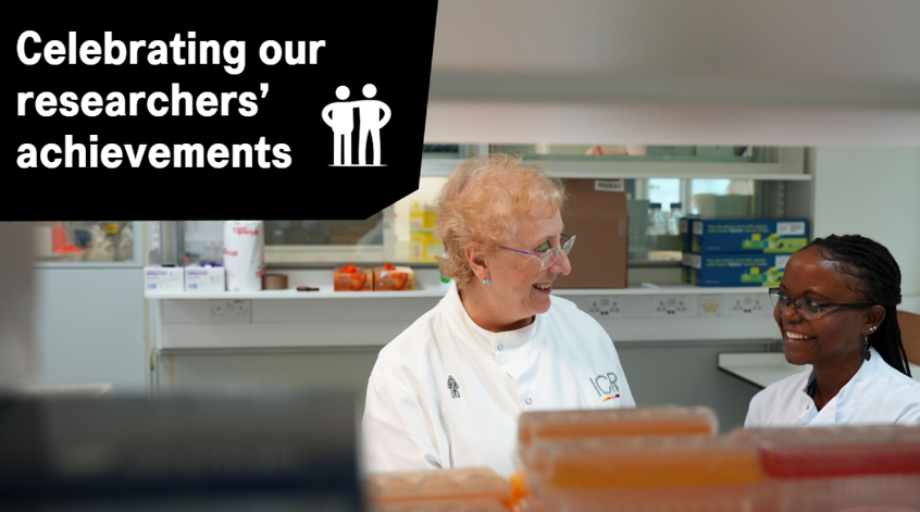 Two female researchers are standing in a lab smiling at each other. One has a man of men badge pinned to her lab coat.