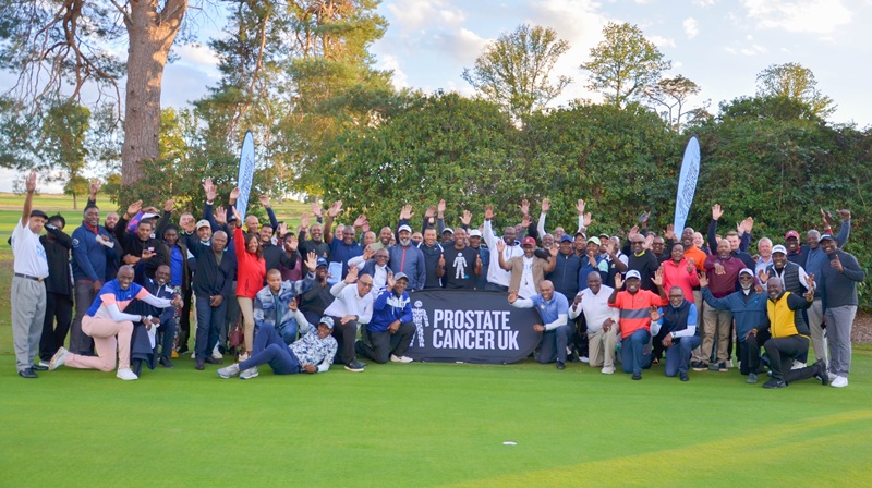A group of people on a golf pitch with a Prostate Cancer UK banner, cheering
