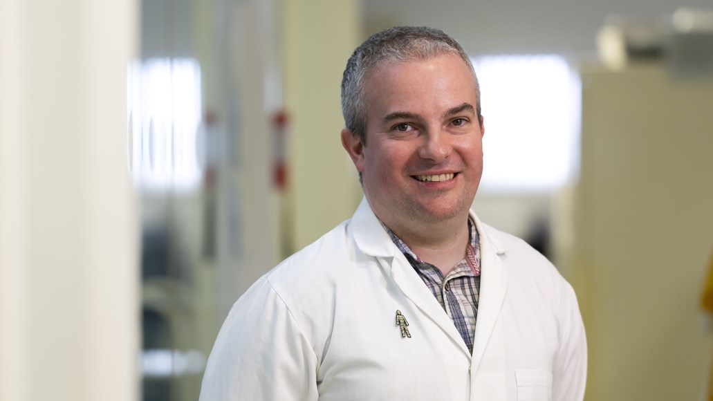 A man with short grey hair wearing a white coat standing in a lab and smiling