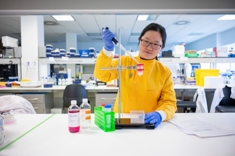 A woman in a lab wearing a yellow lab coat in front of a row of bottles, holding a syringe